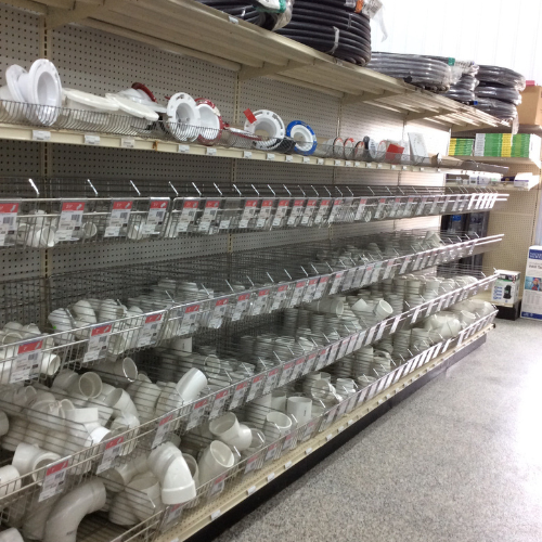 Hardware store aisle with shelves of white plumbing fittings and coiled hoses. Items are neatly arranged in metal baskets. Bright, orderly atmosphere.