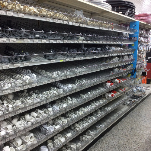 A store aisle with shelves filled with organized plumbing supplies, including white PVC connectors and black hoses. The setting is orderly and well-lit.