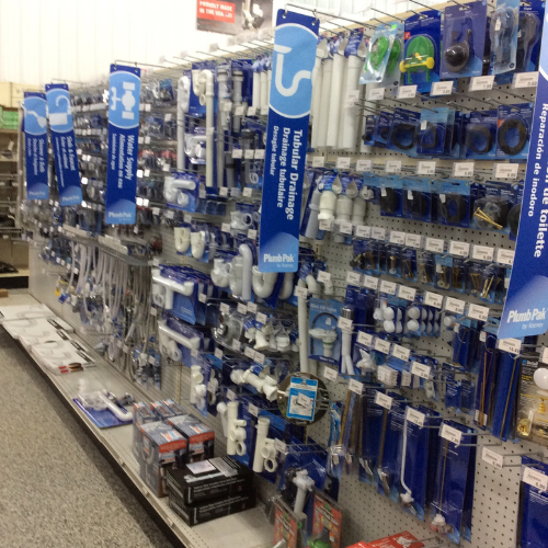 Store aisle with plumbing supplies on metal shelves, featuring blue signs for tubular drainage. Items like pipes and fittings hang neatly, organized, and well-stocked.