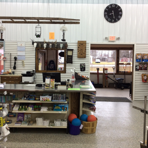 Checkout counter in a general store with office supplies, candy, and sports balls. A black wall clock hangs above, with an open exit door in the background.