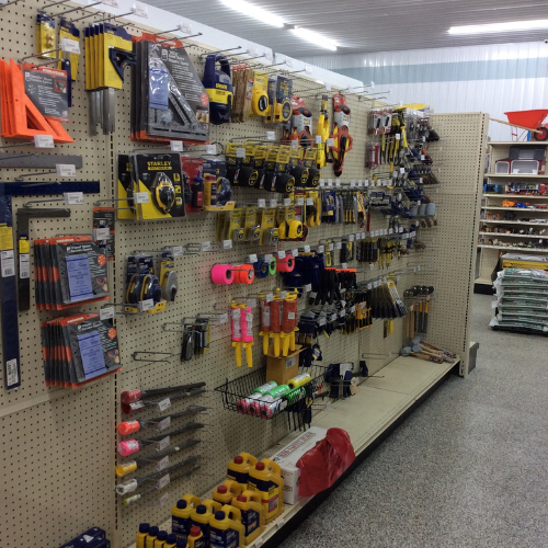Hardware store aisle with neatly organized tools and supplies on pegboard walls. Items include tapes, brushes, and measuring tools, under bright lighting.