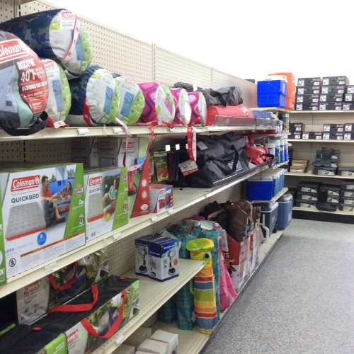 Shelves in a store display neatly stacked camping gear. Items include sleeping bags, air mattresses, coolers, and lanterns, creating an organized and inviting setup.
