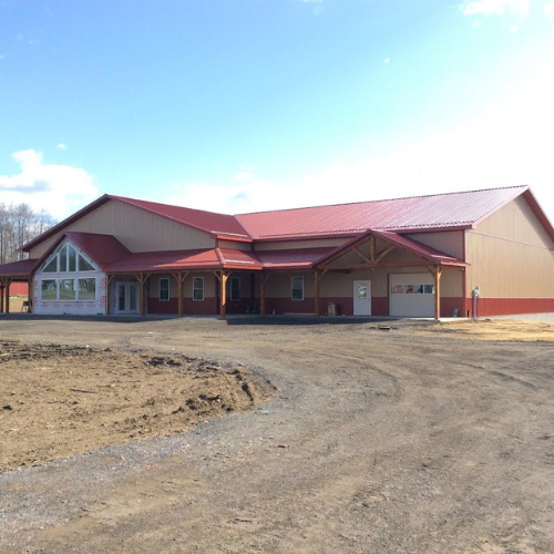 A large beige and red-roofed building stands on a dirt lot under a clear blue sky. The structure features triangular windows and a covered entrance.