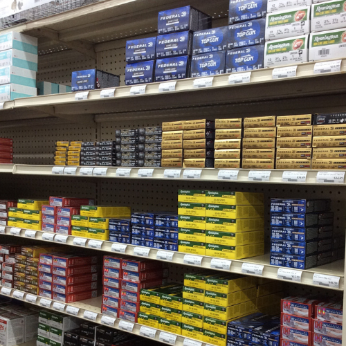 Store shelves filled with neatly stacked boxes of ammunition in various colors, including blue, yellow, and red. Labels display prices and brands.