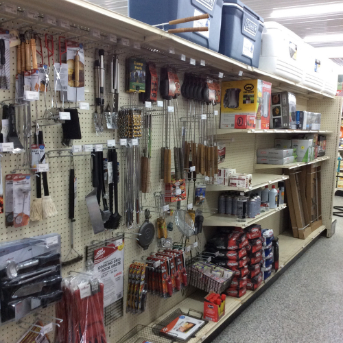 Store aisle with barbecue supplies on display. Shelves hold grilling tools, charcoal, and propane tanks. Coolers are stacked on the top shelf. Bright lighting.