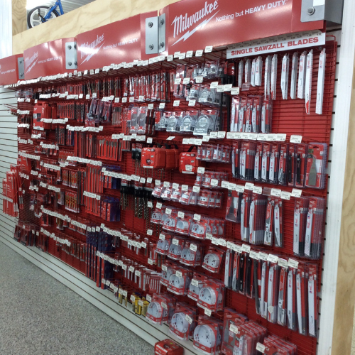 A store display with Milwaukee-branded tools on a red and white slat wall. Items include saw blades and drill bits, neatly arranged. Bright, industrial ambiance.